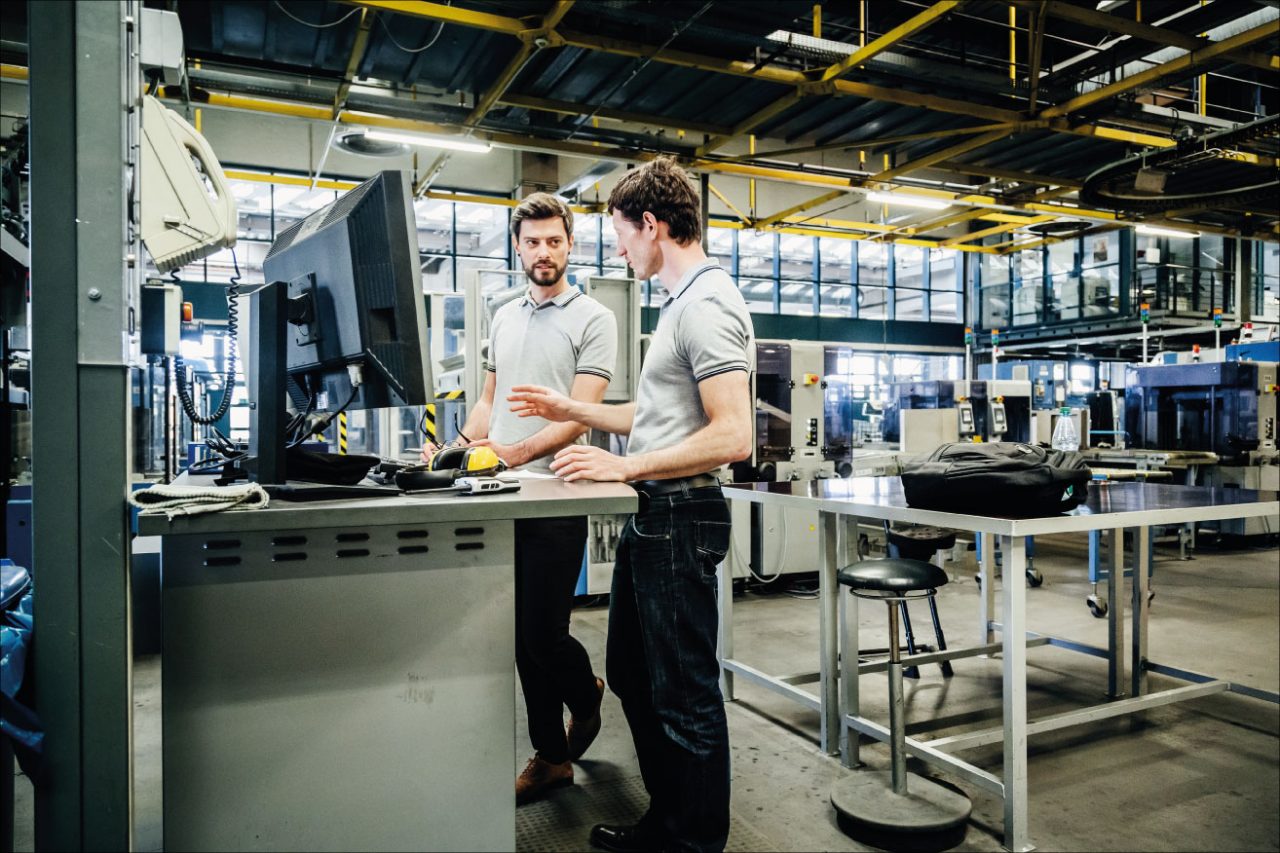 2 men standing in front of a computer in a factory