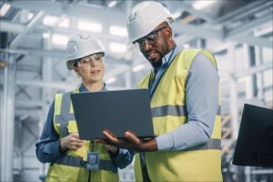 A man and a woman wearing high-visibility vests and safety helmets with a laptop in their hands stand in a production hall